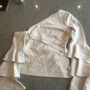 Beige omlottopp med volangärm - Säljer en beige omlottopp med långa ärmar och snygga volangdetaljer. Toppen har oneshoulder-design och knytband i midjan för en cool siluett. Materialet känns mjukt och har en lätt glans. Dragkedja i sidan för enkel påklädning. Köpt i UK .