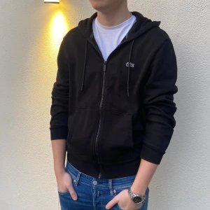 Svart hoodie från Lacoste med dragkedja - Svart hoodie från Lacoste i storlek Tillverkad i mjuk bomull och har ribbade muddar vid ärmslut och nederkant. Perfekt för chill dagar.
