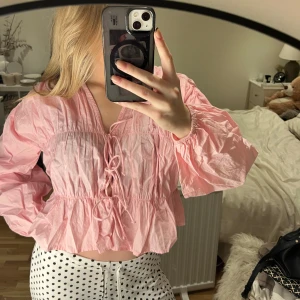 Rosa blus med snörning och volang - Säljer en ljusrosa blus med v-ringning och snörning framtill. Blusen har långa puffiga ärmar, volang nedtill och är croppad i modellen. Materialet är lätt och krispigt, perfekt för vår och sommar. Superfin att styla med jeans eller kjol!