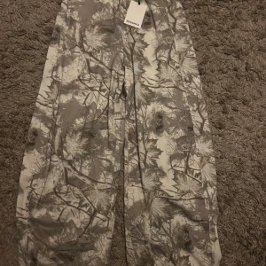 Camouflage cargopants från Bershka - Säljer ett par gråa och beige cargopants från Bershka med snyggt camouflage-mönster. Byxorna har en loose passform och raka ben, perfekta för dig som gillar streetstyle. Materialet är mjukt och bekvämt, och midjan är normal. Taggen sitter kvar.