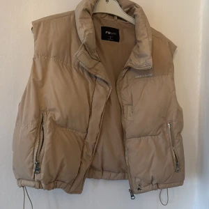Beige dunväst från FB Sister - Beige dunväst från FB Sister med hög krage, dragkedja framtill och två fickor med dragkedja. Västen har justerbar dragsko nertill och broderad text på bröstet. Perfekt för lager-på-lager och chill stil.