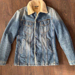 Blå jeansjacka Nudie Jeans - Säljer en snygg blå jeansjacka från Nudie Jeans med mjuk teddykrage och foder. Jackan har klassiska bröstfickor med knappar, raka sidofickor och coola kontrastsömmar. Perfekt för dig som gillar streetstyle och vill ha en varm jacka med vintagekänsla.Nypris runt 2500. Hör av dig vid frågor!!!