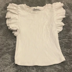 Vit ribbad topp med volangärmar från Zara - Supersöt vit topp från Zara i ribbad bomull med korta volangärmar som ger en extra touch. Rund halsringning och figurnära passform. Perfekt för dig som gillar stilrena och feminina detaljer.