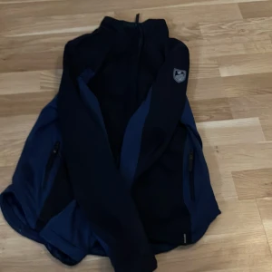 Mörkblå vindjacka med dragkedja 153-162 - Snygg mörkblå vindjacka från Craft med hög krage och hel dragkedja framtill. Jackan har två sidofickor med dragkedja och sportig design med blåa paneler längs sidorna. Perfekt för dig som gillar en clean och funktionell look.