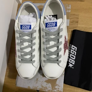 GGDB vita sneakers med rosa glitterstjärna - Snygga vita sneakers från Golden Goose med grå snörning, ljusblå häl och en glittrig rosa stjärna på sidan. Skorna har en klassisk rund tå och platt sula, perfekt för dig som vill sticka ut med en cool detalj. Tillverkade i skinn och med GGDB-logga på plösen.