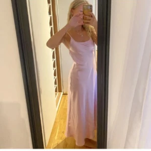 Ljusrosa långklänning i satin - Superfin ljusrosa långklänning i satin med smala axelband och öppen rygg. Klänningen har en mjukt fallande siluett och en enkel, elegant look som känns både trendig och tidlös. Perfekt för dig som gillar minimalistisk stil och vill sticka ut med en feminin touch.