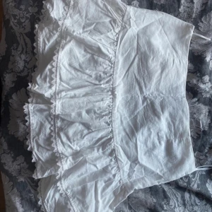 Vit volangkjol med spetsdetaljer - Säljer en söt vit kortkjol med volanger och spetsdetaljer längs kanterna. Kjolen har en luftig och lätt känsla, perfekt för varma dagar. Materialet är mjukt och känns bekvämt mot huden. Volangerna ger kjolen en extra feminin touch.