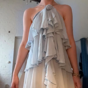 Ljusblå långklänning med volanger - Säljer en ljusblå långklänning med flera lager volanger och en söt rosett vid halsen. Klänningen har halterneck och är ärmlös, vilket ger en luftig och flowy känsla. Materialet är tunt och skönt, perfekt för varma dagar. Dragkedja i ryggen för enkel påklädning.