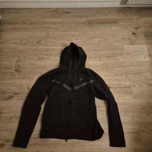 Svart Nike hoodie med dragkedja M - Svart hoodie från Nike i storlek M med hel dragkedja framtill och huva. Snygga svarta detaljer i form av två diagonala ränder över bröstet. Långärmad och tillverkad i mjukt material som passar perfekt för chill eller träning.