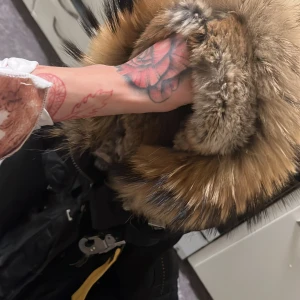 Svart Parajumpers dunjacka med päls - Säljer en svart Parajumpers dunjacka med stor fluffig päls runt huvan och coolt gult spänne vid kragen. Jackan har patch på ärmen, flera fickor och dragkedja framtill. Perfekt för dig som vill ha en varm och snygg vinterjacka med street vibe.