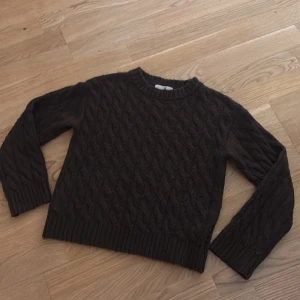 Mörkbrun stickad tröja med kabelmönster - Snygg mörkbrun stickad tröja med klassiskt kabelmönster och rund hals. Tröjan har långa ärmar och ribbade muddar vid hals, ärmslut och nederkant. Perfekt för kyliga dagar och passar till jeans eller kjol.