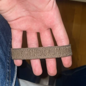 Christian Dior J'Adior armband - Snyggt armband från Christian Dior med broderad text 'Christian Dior J'Adior' i beige-brun ton. Armbandet är gjort i tyg och har justerbar knytning för perfekt passform. Perfekt accessoar för dig som gillar designer och vill ha något unikt på handleden.