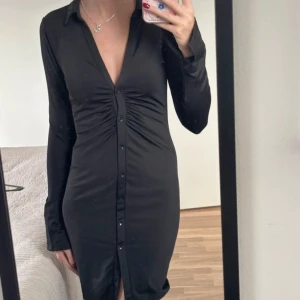 Svart kort klänning med knappar - Svart kort klänning med lång ärm och djup v-ringning. Klänningen har knappar hela vägen framtill och snygg rynkdetalj vid bysten. Figurnära passform och mjukt, stretchigt material som känns bekvämt. Perfekt för dig som gillar stilrena och trendiga plagg.