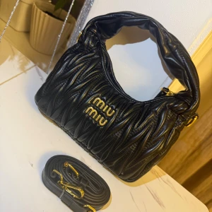 Svart quiltad axelväska  - Snygg svart axelväska  i quiltat skinn med guldiga detaljer och logotyp framtill. Väskan har en mjuk, rundad form och medföljande axelrem med guldfärgade spännen. Perfekt för dig som gillar lyxiga accessoarer med cool vibe.