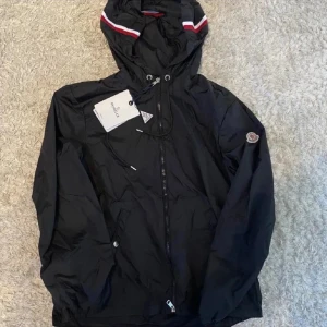 Svart vindjacka från Moncler - Svart vindjacka från Moncler med huva och snörning, dragkedja framtill och Moncler-logga på ärmen. Jackan har vita och röda detaljer på huvan och praktiska fickor med knappstängning. Perfekt för dig som gillar stilren och sportig look.