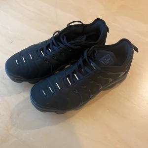 Nike Air Vapormax Plus svarta sneakers - Svarta Nike Air Vapormax Plus sneakers med futuristisk design och synlig luftdämpning i sulan. Skorna har snörning, diskreta reflexdetaljer och en chunky, genomskinlig sula. Ovandelen är i syntetmaterial och mesh för en sportig vibe.
