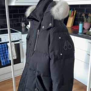 Svart dunjacka med päls från Moose Knuckles - Säljer en svart dunjacka från Moose Knuckles med fluffig vit päls på huvan. Jackan har flera dragkedjor, knappar och ett snyggt logomärke på ärmen. Köpt 2022 men knappt använd då jag känt att jag ej passar. Köpt från Johnells 