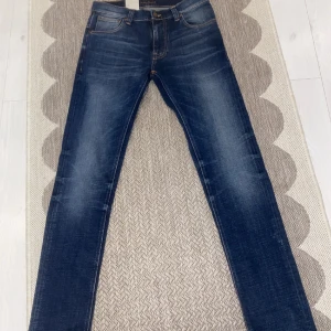 Blå jeans från Nudie - Snygga blå jeans från Nudie med klassisk femficksdesign kontrastsömmar. och har en mörk tvätt med lätt slitning på låren och knäna. Materialet är mjukt denim i bomull och jeans, perfekt för en cool och avslappnad look. Dem är enbart testade alla lappar och allt finns kvar på jeansen 