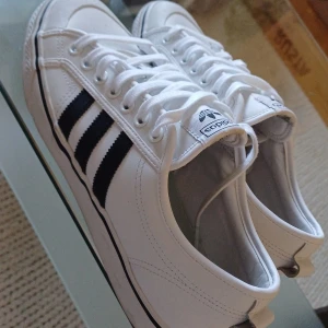 Adidas Nizza vita sneakers med svarta ränder - Säljer ett par fräscha Adidas Nizza sneakers i vitt med klassiska svarta ränder på sidorna. Skorna har snörning, platt sula och är tillverkade i syntetmaterial och textil. Perfekta för dig som gillar retrovibbar och stilren design.