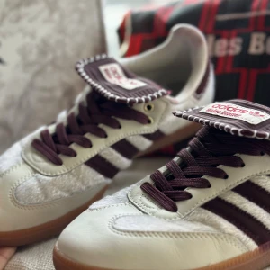 Adidas Wales Bonner Samba sneakers - Unika Adidas Wales Bonner Samba sneakers i vitt och mörkbrunt med klassiska tre ränder och kontrasterande sula. Skorna har detaljer i skinn och mocka, grova mörkbruna snören och specialdesignad plös med vit dekorationssöm. Perfekt för dig som vill sticka ut med din stil.Size 39 1/2