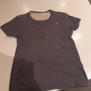 Mörkgrå t-shirt från Guess - Snygg mörkgrå t-shirt från Guess med tryck på ryggen och liten röd logga framtill. Klassisk rund halsringning och korta ärmar. Perfekt för dig som gillar streetwear och vill ha något med cool LA-vibe.