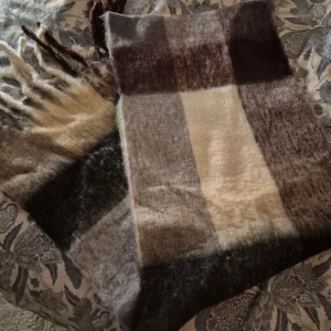 Rutig fluffig halsduk i jordnära toner - Stor och supermjuk halsduk med rutigt mönster i brunt, beige, grått och svart. Lång modell med rejäla fransar i ändarna, perfekt att svepa runt sig för extra värme. Ger en mysig och trendig vibe till din outfit.