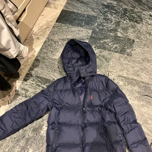 Ralph Lauren Jacka - Snygg mörkblå pufferjacka från Polo Ralph Lauren med huva och dragkedja. Jackan har det klassiska röda Polo-loggan broderad på bröstet och två fickor med dragkedja. Perfekt för kalla dagar när du vill hålla stilen.