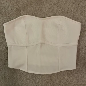 Vit ribbad bandeau tubtopp - Säljer en stilren vit bandeau tubtopp med ribbad struktur och snygga sömmar som ger en korsettliknande känsla. Toppen är axelbandslös och har en lätt böjd kant upptill för en schysst passform. Perfekt att styla med jeans eller kjol.