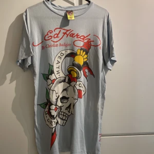 Ed Hardy t-shirt med döskalletryck - Ljusblå t-shirt från Ed Hardy med stort döskalletryck och texten 'Death or Glory' framtill. Röd Ed Hardy-logga både fram och bak. Klassisk rund hals och korta ärmar. Materialet är mjuk bomull och passformen är normal. Perfekt för dig som gillar streetwear och edgy motiv. Några få små hål.