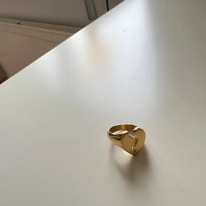 Guldfärgad ring med hjärtdetalj - Cool guldfärgad ring med ett hjärta som har en blixtformad spricka i mitten. Ringen har en chunky design och glänsande finish som ger den en unik och edgy vibe. Perfekt för dig som gillar statement-smycken med lite attityd.