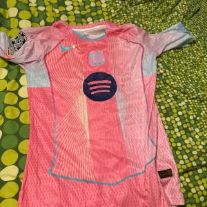 Rosa FC Barcelona fotbollströja Nike - Säljer en riktigt snygg FC Barcelona fotbollströja från Nike i rosa och ljusblå toner. Tröjan har korta ärmar, coolt grafiskt mönster och Spotify-logga på bröstet. Perfekt för dig som vill sticka ut på planen eller supporta Barca.