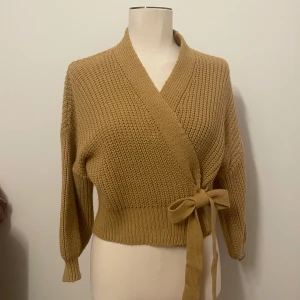 Beige stickad omlottkofta med knyt - Superfin beige stickad omlottkofta med knyt i midjan och bred ribbad kant. Koftan har lång ärm och en mysig, chunky känsla som passar perfekt till lager-på-lager. Enkel att styla och riktigt trendig färg för säsongen.