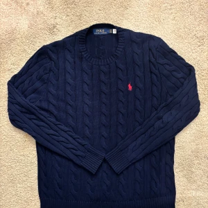 Mörkblå stickad tröja Polo Ralph Lauren - Kabelstickad mörkblå tröja från Polo Ralph Lauren med klassisk rund hals och röd broderad logga på bröstet. Tröjan har långa ärmar och ribbade muddar, perfekt för lager-på-lager under kyliga dagar. Använt ett få par gånger! 2-3 gånger snackar vi om 😀