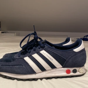 Adidas L.A. Trainer sneakers marinblå - Säljer ett par Adidas L.A. Trainer sneakers i marinblått och vitt. Skorna har klassiska tre ränder på sidan, snörning och detaljer i mocka och mesh. Sulan har de ikoniska pluggarna i rött, vitt och svart. Perfekta för dig som gillar retrostil och sportig vibe.