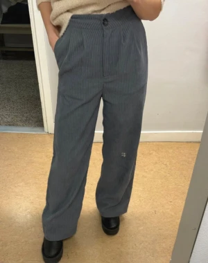 Gråa randiga manchesterbyxor wide - Snygga gråa manchesterbyxor med diskret randigt mönster och breda ben. Byxorna har hög midja, klassisk knappstängning och sidofickor. Perfekt för dig som gillar en chill och trendig look med lite retrovibe.