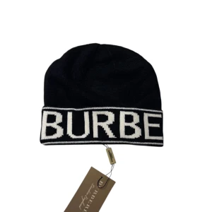 Svart Burberry mössa med logga - Svart mössa från Burberry med bred vit uppvikt kant där BURBERRY är stickat i stora bokstäver runt hela mössan. Klassisk och stilren design, perfekt för dig som gillar exklusiva märken. Materialet är stickad ull som känns mjuk och varm.
