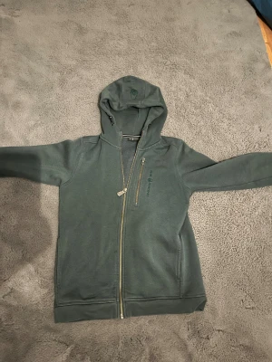 Grön hoodie från Sail Racing med dragkedja - Mörkgrön hoodie från Sail Racing med broderad logga på bröstet och huvan. Tröjan har hel dragkedja framtill, en extra bröstficka med dragkedja och mjukt fleecefoder på insidan. Klassisk passform och långa ärmar, perfekt för chilliga dagar.