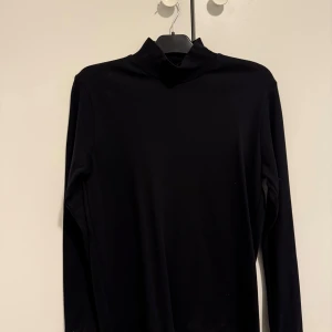 Svart polotröja från Clipper - Svart polotröja från Clipper i storlek S. Tröjan har en klassisk hög krage och är långärmad med mjukt material som känns skönt mot huden. Perfekt att styla med jeans eller under en jacka. Enkel och stilren design utan mönster eller detaljer.