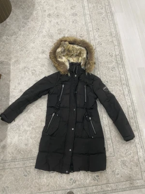 Svart Rockandblue dunjacka med äkta päls - Lång svart dunjacka från Rockandblue med snygg äkta päls runt huvan. Jackan har flera dragkedjor, tryckknappar och coola detaljer på ärmen. Perfekt för kalla vinterdagar och har en riktigt urban vibe. Äkta päls inuti luvan med.