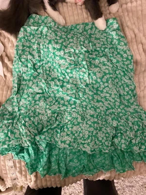 Grön blommig volangkjol från Pieces/hm - Snygg kort kjol från Pieces i en fräsch grön färg med vita blommor. Kjolen har volangdetaljer och ett smockat midjeband som ger en skön passform. Perfekt för dig som gillar färg och mönster! Materialet är mjukt och lätt, vilket gör den superbekväm.