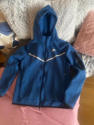 Blå Nike kofta med huva - Snygg blå vindjacka från Nike med huva och hel dragkedja framtill. Jackan har en vit detalj över bröstet och en ficka med dragkedja på ärmen. Perfekt för dig som gillar sportig stil och vill ha något lätt och smidigt.