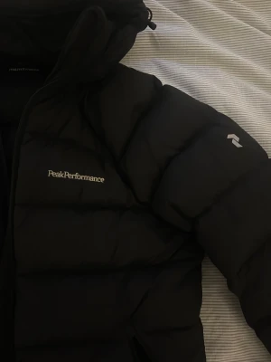 Svart dunjacka Peak Performance - Svart dunjacka från Peak Performance med huva och dragkedja framtill. Jackan har en puffig look, två fickor med dragkedja och diskret logga på bröstet samt ärmen. Perfekt för kalla dagar och riktigt snygg till streetwear.