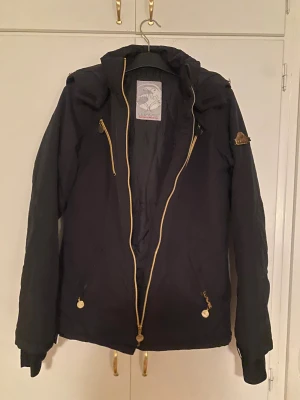 Svart vinterjacka för ungdom från Wave Sportwear - Svart vinterjacka från Wave Sportwear till ungdom med guldiga dragkedjor och detaljer. Strl S/M. Jackan har huva, hög krage och flera fickor med dragkedja. Perfekt för kalla dagar och har en snygg, sportig look. Materialet är syntet och jackan är långärmad.