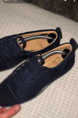 Mörkblå sneakers från Christian Louboutin - Snygga mörkblå sneakers från Christian Louboutin i mocka med klassisk rund tå och snörning. Insidan är beige och yttersulan har den ikoniska röda färgen med Louboutin-logga. Perfekt för dig som vill sticka ut med exklusiva detaljer och stilren design. Allt ox följer med och självklart äkta
