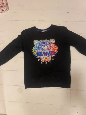 Svart Kenzo sweatshirt - Svart sweatshirt från Kenzo med ikonisk broderad tiger i blått, rött, gult och silver på bröstet. Tröjan har rund hals, långa ärmar och ribbade muddar. Perfekt statement-plagg med färgstark design. Passar som s och m