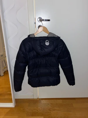 Marinblå pufferjacka från Sail Racing - Marinblå pufferjacka från Sail Racing med huva och grått innerfoder. Jackan har dragkedja framtill och logga på bröstet samt en snygg detalj på ryggen. Perfekt för kalla dagar när du vill hålla stilen och värmen.