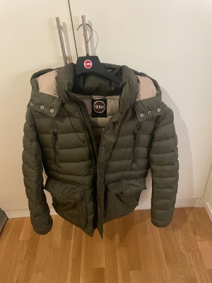 Olivgrön  Colmar Jacka  - Snygg olivgrön pufferjacka från Colmar med huva och flera praktiska fickor med dragkedja och knappar. Jackan har quiltad design och är fodrad i ljusare beige. Perfekt för vinern och har en modern, sportig look.