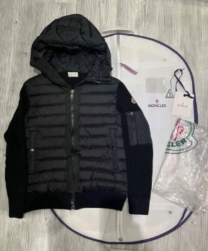 Svart Moncler cardigan med huva - Snygg svart pufferjacka från Moncler med stickade ärmar och vadderad kropp. Jackan har huva, dragkedja framtill, fickor med dragkedja och Moncler-logga på ärmen. Perfekt för kyliga dagar och ger en clean, exklusiv look.