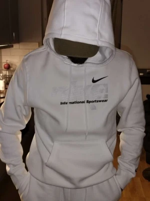 Vit Nike hoodie med tryck och huva - Säljer en clean vit Nike hoodie med svart logga och texten 'International Sportswear' på bröstet. Hoodien har huva med snörning, känguruficka och tryck även på ryggen. Mjuk insida och relaxed fit, perfekt för chill dagar. En helt ny dress storlek smal men passar verkligen medium om inte l också då de e rätt stora i storlek. Ordinarie pris hoddien 799 mjukisen 749 . Släpper för 1200  som sagt helt ny endast testad hemma 