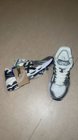 ASICS sneakers i silver och vitt - Säljer ett par snygga ASICS sneakers i silver, vitt och svart med coola detaljer. Skorna har mesh-överdel för bra ventilation, silvriga paneler och klassisk snörning. Sulan är vit med svarta inslag och har en sportig design som sticker ut. Perfekta för dig som gillar retrostil och komfort.